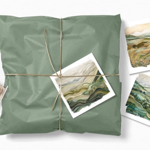May include: A set of three landscape art prints featuring abstract watercolor paintings of rolling hills and valleys in shades of green, brown, and beige. The prints are packaged in a green paper bag tied with twine.
