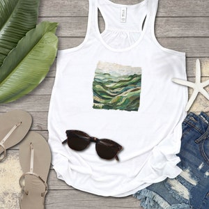 May include: White tank top with a green and blue mountain landscape graphic print. The tank top is laying on a wooden surface with a pair of sandals, sunglasses, and denim shorts.