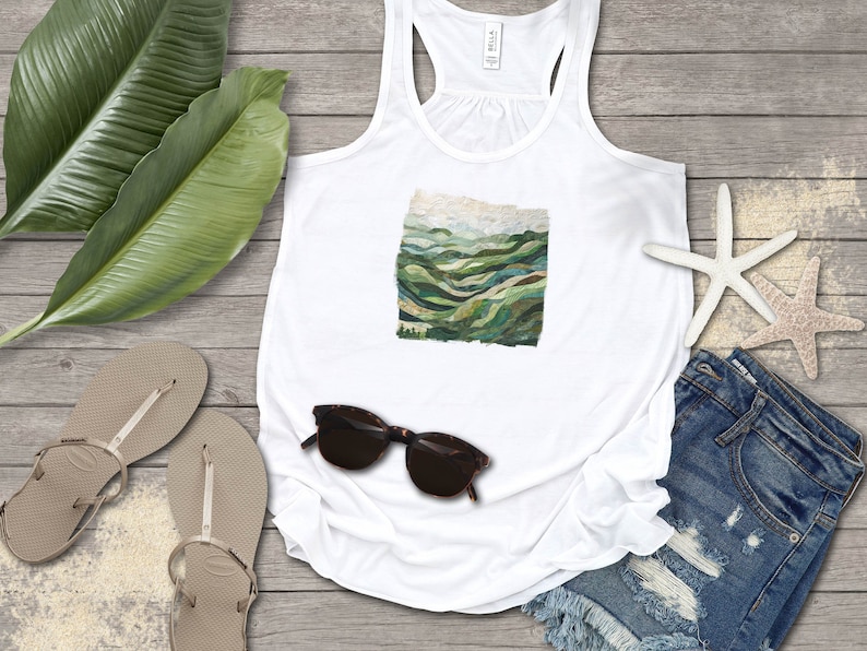 May include: White tank top with a green and blue mountain landscape graphic print. The tank top is laying on a wooden surface with a pair of sandals, sunglasses, and denim shorts.