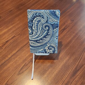 May include: A book cover with a blue and white paisley pattern. The cover is made of fabric and has a white bookmark. The book cover is on a wooden surface.