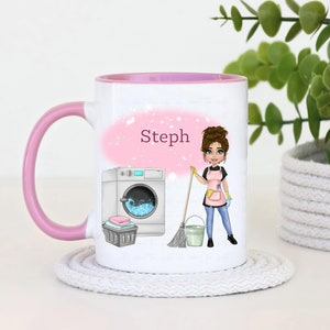Könnte beinhalten: Ein weißer Keramikbecher mit rosa Henkel und Rand, verziert mit einer Cartoon-Illustration einer Person mit braunen Haaren in einer rosa Schürze, die einen Besen neben einem Eimer, einer Waschmaschine, einem Wäschekorb und dem Namen Steph hält.