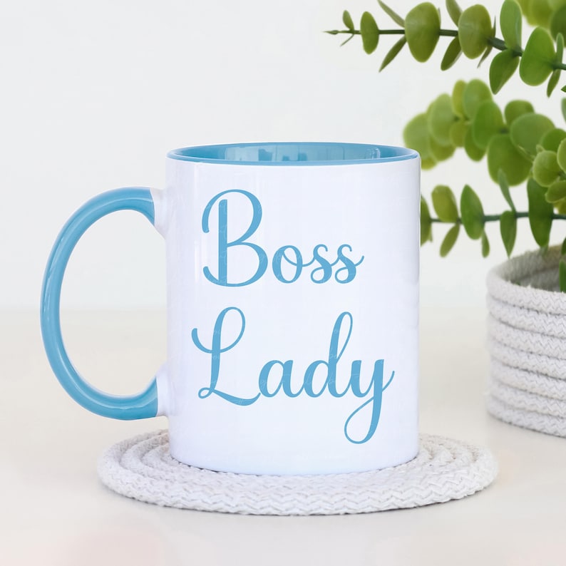 Puede incluir: Taza de cer&aacute;mica blanca con interior y asa azul claro. La taza presenta las palabras "Boss Lady" en escritura azul claro. La taza se encuentra sobre un posavasos circular blanco. Una planta en maceta est&aacute; en el fondo.