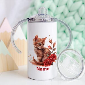 Peut inclure: Un gobelet en acier inoxydable blanc avec un motif de chaton et de roses rouges. Le gobelet a deux poignées et un couvercle transparent. Le texte "Name" est imprimé sur le gobelet.