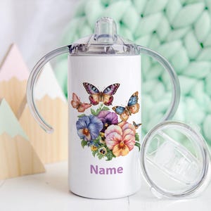 Peut inclure: Gobelet à bec blanc avec un motif de papillons et de fleurs. Le gobelet a un couvercle et des poignées transparents. Le motif comprend des papillons aux tons orange, bleu et rose, avec des fleurs violettes et orange. Le mot "Name" est imprimé sur le gobelet.