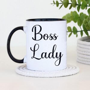 Puede incluir: Taza de cer&aacute;mica blanca con interior y asa negros. La taza presenta las palabras "Boss Lady" en una fuente negra. Se apoya en un posavasos redondo blanco. Una planta verde est&aacute; en el fondo.