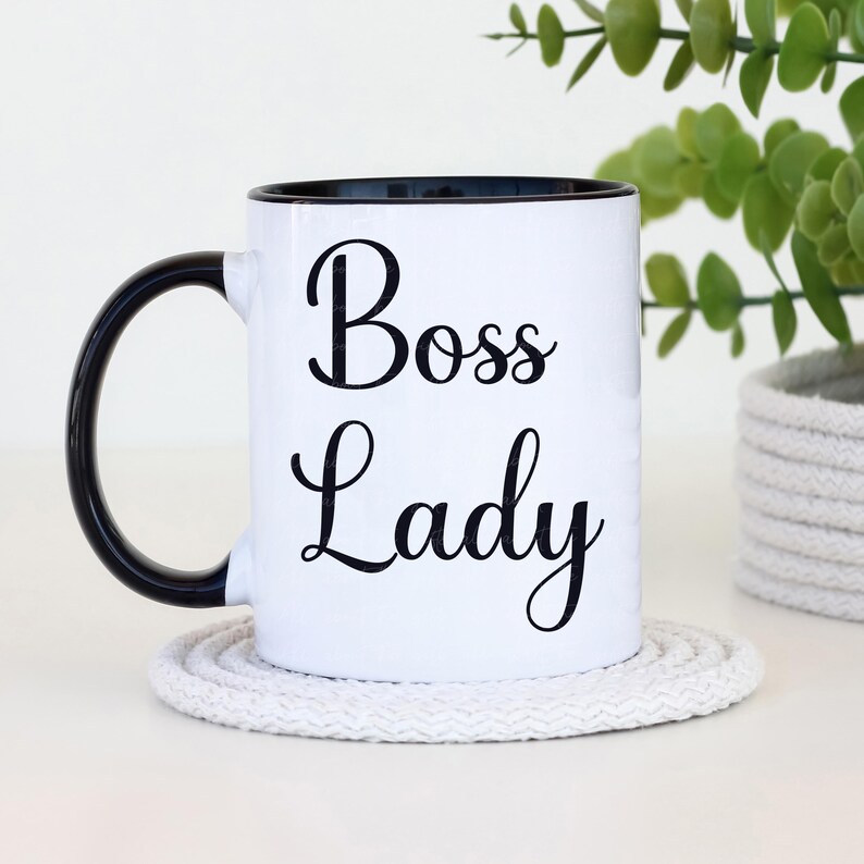 Puede incluir: Taza de cer&aacute;mica blanca con interior y asa negros. La taza presenta las palabras "Boss Lady" en una fuente negra. Se apoya en un posavasos redondo blanco. Una planta verde est&aacute; en el fondo.