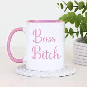 Puede incluir: Taza de cer&aacute;mica blanca con interior y asa rosas. La taza presenta las palabras "Boss Bitch" en rosa. La taza est&aacute; sobre un posavasos blanco, con una planta verde en el fondo.