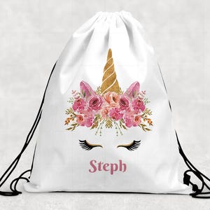 May include: A white drawstring backpack featuring a gold glitter unicorn horn, pink floral crown, and black eyelashes. The name Steph is printed in pink lettering below the eyelashes. The bag has black cord straps on both sides for closure.