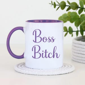 Puede incluir: Taza de cer&aacute;mica blanca con interior y asa morados. La taza presenta las palabras "Boss Bitch" en escritura morada. La taza est&aacute; sobre un posavasos blanco y circular. Una peque&ntilde;a planta en una maceta tejida est&aacute; en el fondo.