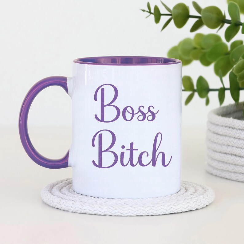 Puede incluir: Taza de cer&aacute;mica blanca con interior y asa morados. La taza presenta las palabras "Boss Bitch" en escritura morada. La taza est&aacute; sobre un posavasos blanco y circular. Una peque&ntilde;a planta en una maceta tejida est&aacute; en el fondo.