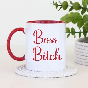 Puede incluir: Taza de cer&aacute;mica blanca con interior y asa rojos. La taza presenta las palabras "Boss Bitch" en escritura roja. La taza est&aacute; sobre un posavasos blanco y circular.