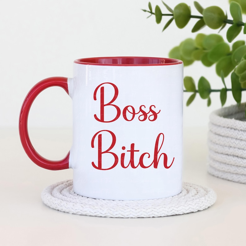 Puede incluir: Taza de cer&aacute;mica blanca con interior y asa rojos. La taza presenta las palabras "Boss Bitch" en escritura roja. La taza est&aacute; sobre un posavasos blanco y circular.