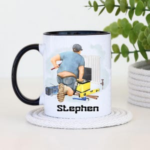 Puede incluir: Taza de cerámica blanca con asa y borde negros. La taza presenta una ilustración de dibujos animados de un fontanero en el trabajo, con el nombre "Stephen" impreso en negro. La taza está sobre un posavasos blanco.