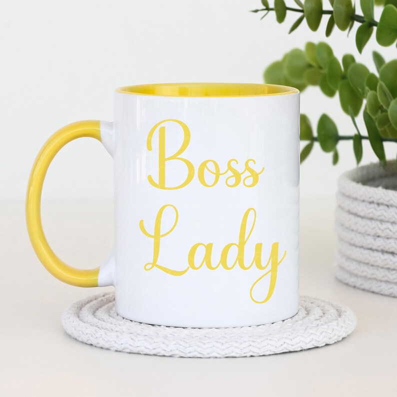 Puede incluir: Taza de cer&aacute;mica blanca con interior y asa amarillos. La taza presenta las palabras "Boss Lady" en escritura amarilla. Se apoya en un posavasos blanco tejido. Una planta verde en una maceta gris est&aacute; en el fondo.