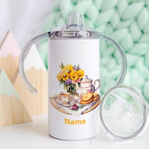 Può includere: Un bicchiere in acciaio inossidabile bianco con un design floreale e la parola "Name" stampata sul fondo. Il bicchiere ha due manici e un coperchio trasparente.