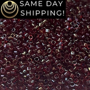 May include: A close-up image of a large quantity of small, dark red, iridescent glass seed beads. The beads are arranged in a random pattern and have a shiny, metallic finish.