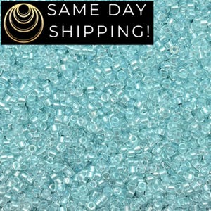 May include: A close-up shot of a pile of small, light blue seed beads. The beads are cylindrical with a slightly translucent appearance. The image has a soft focus, highlighting the texture and color of the beads. The text "SAME DAY SHIPPING!" is in the top left corner.