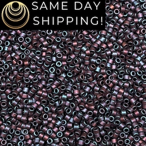 May include: A large collection of small, cylindrical seed beads with a metallic, iridescent finish that shifts between deep purple, bronze, and dark blue tones. The beads are uniform in shape and size, creating a dense, textured surface.