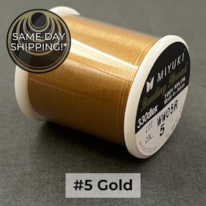 May include: A spool of gold-colored Miyuki beading thread made of 100% nylon. The label on the white plastic spool indicates 330dtex, lot number WW05R, and color number 5. The thread is wound tightly around the spool, which is labeled #5 Gold.
