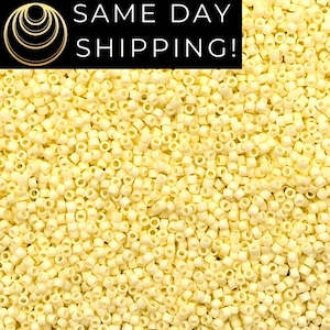 May include: A large collection of small, cylindrical, pale yellow beads with a matte finish. Each bead features a central hole, creating a uniform, textured appearance across the entire surface. The beads are densely packed together in a pile.