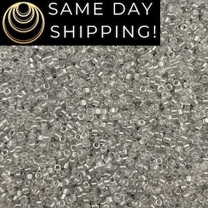 May include: A close-up image of a large quantity of small, clear glass seed beads. The beads are round and have a slight iridescent sheen.