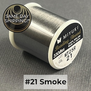 May include: A spool of dark grey Miyuki beading thread made of 100% nylon. The label displays the text Miyuki Beading Thread, 330dtex, Made in Japan, Lot WT25R, and Col 21. The thread is a smoke color and is wound around a white plastic spool.