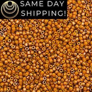 May include: A large collection of small, cylindrical glass beads in a warm, metallic amber-orange hue. Each bead features a consistent tubular shape with a central hole, displaying a glossy, reflective finish across the entire surface.