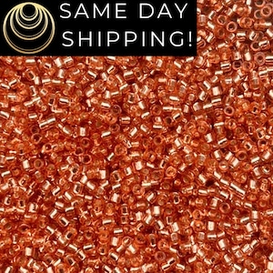 May include: Close-up of a pile of small, copper-colored seed beads. The beads are cylindrical with rounded edges, reflecting light. The image includes the text "SAME DAY SHIPPING!" in white letters on a black background.