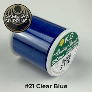 May include: A spool of K.O. Beading Thread in a clear blue color. The spool features a green label with white text reading 330dtex, 100% nylon, made in Japan, and includes lot number IN20R and color code 21CB. The thread is wound tightly on the spool.