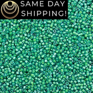 May include: A close-up shot of a pile of small, cylindrical, green seed beads. The beads are a mix of light and dark green, creating a textured appearance. The image includes the text "SAME DAY SHIPPING!" in white letters on a black background.