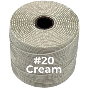 May include: A cylindrical spool of cream-colored twisted cord featuring a dark brown center core. The text #20 Cream is printed in white on the front of the spool, indicating the specific size and color of the cord wound tightly around the spool.
