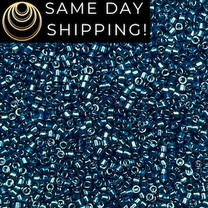 May include: A large collection of small, cylindrical, metallic blue glass beads with a reflective finish. The beads are densely packed together, showing their uniform shape and consistent deep blue color throughout the entire frame.