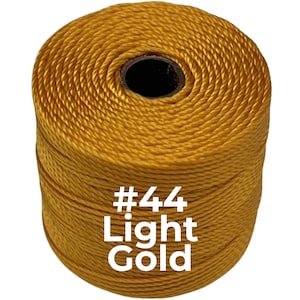 May include: A cylindrical spool of twisted cord in a light gold color. The spool features a dark center core and is labeled with the white text #44 Light Gold printed directly onto the front of the golden cord surface.