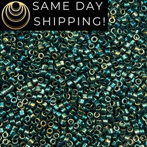May include: A large collection of small, cylindrical seed beads with a metallic, iridescent teal and dark blue finish. The beads are densely packed together, showing a reflective, glossy surface texture with varying shades of deep ocean blue.