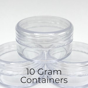 May include: Three clear, round, empty plastic jars stacked in a pyramid formation. Each container features a screw-top lid and a smooth, transparent finish. The text 10 Gram Containers is printed in black on the front of the bottom two jars.