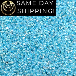 May include: A close-up shot showcases a sea of light blue seed beads. The beads are small, cylindrical, and have a slightly iridescent sheen. The image is filled with the beads, creating a textured, shimmering effect. The text "SAME DAY SHIPPING!" is at the top.