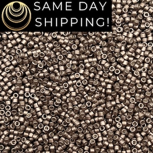 May include: A close-up image of a large quantity of small, round, metallic beads. The beads are a light brown color with a slightly metallic sheen. The beads are arranged in a random pattern, creating a textured surface.