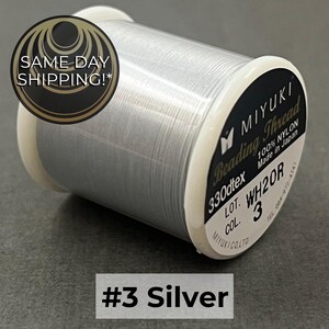 May include: A spool of silver Miyuki beading thread made of 100% nylon. The label on the white plastic spool indicates 330dtex, lot WH20R, and color number 3. The thread is wound tightly around the spool, creating a smooth, metallic appearance.
