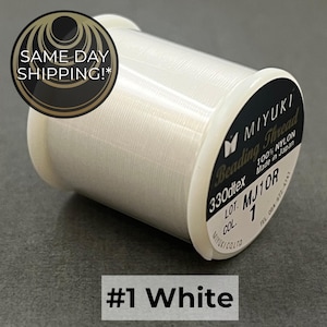 May include: A spool of white Miyuki beading thread made of 100% nylon. The label on the spool indicates 330dtex, lot MJ10R, and color 1. The spool is white with a black circular label on the side containing white text and branding details.