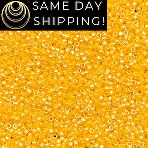 May include: Close-up of a pile of small, yellow seed beads. The beads are cylindrical with a hole running through the center. The image has a bright, sunny appearance, suggesting a craft or jewelry-making supply.
