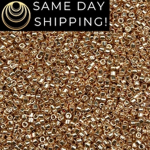 May include: A close-up image of a large quantity of small, gold-colored seed beads. The beads are round and have a metallic sheen.