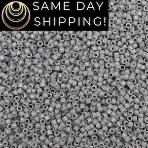 May include: A large collection of small, cylindrical, opaque light gray beads with a smooth finish. The beads are uniform in shape and size, featuring a central hole, and are densely packed together to create a consistent, textured surface.