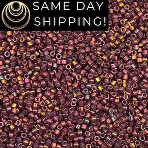May include: A large collection of small, cylindrical glass beads with a metallic, iridescent plum-purple finish. Each bead features a central hole and a reflective surface that catches light, creating a dense, uniform texture across the frame.