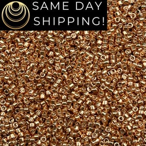 May include: A close-up image of a large quantity of small, gold-colored seed beads. The beads are round and have a metallic sheen.