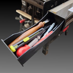 May include: A black tool drawer with a variety of tools inside, including a wooden handled carving knife, a utility knife with a green blade, a red handled screwdriver, and a few pens.