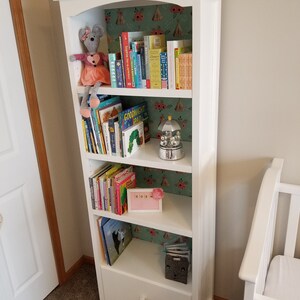 Op de afbeelding: Een witte boekenkast met vijf planken vol kinderboeken. De achterkant van de boekenkast is bekleed met een groen en roze patroonpapier. Een knuffelmuis zit op de bovenste plank. Een klein roze fotolijstje met een babyfoto staat bovenop de boekenkast.