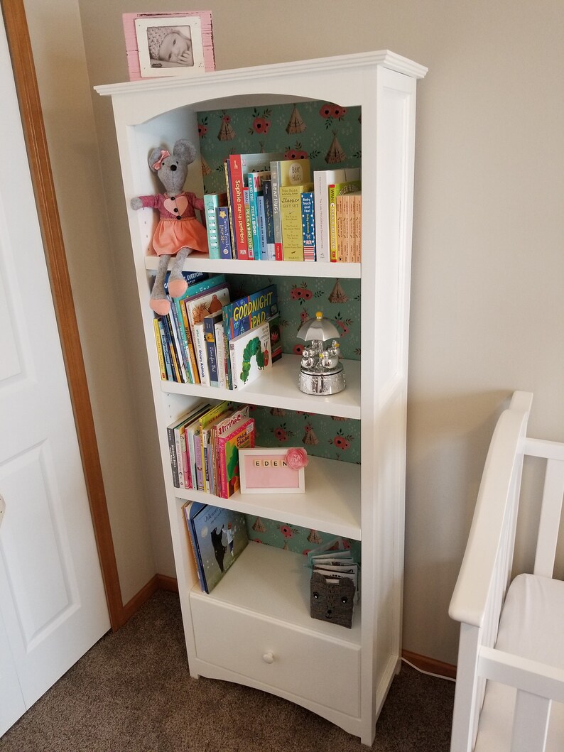 Op de afbeelding: Een witte boekenkast met vijf planken vol kinderboeken. De achterkant van de boekenkast is bekleed met een groen en roze patroonpapier. Een knuffelmuis zit op de bovenste plank. Een klein roze fotolijstje met een babyfoto staat bovenop de boekenkast.