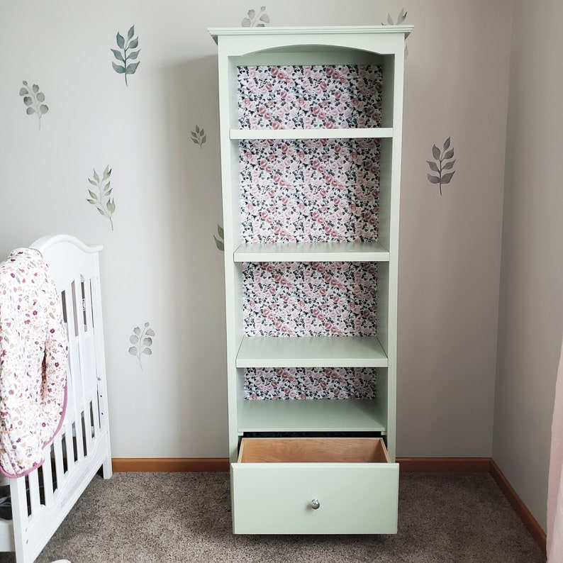 Op de afbeelding: Een lichtgroen boekenkast met vier planken en een lade. De planken zijn bekleed met behang met bloemenmotief. De boekenkast staat op een lichtbruine vloer.