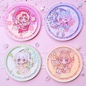 May include: Four round resin coasters with glitter and cartoon character designs. Each coaster features a different character on a crescent moon, with pastel pink, purple, green, and yellow colour schemes. The text "wingmadewithlove" is at the top.