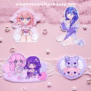 Peut inclure: Quatre autocollants représentant des personnages de style anime avec des couleurs pastel et un fond rose. Les autocollants représentent une fille jouant à des jeux vidéo avec un petit animal, une fille aux cheveux bleus et une veste violette, deux filles jouant à des jeux vidéo ensemble et deux petits animaux dans un cadre en forme de cœur.